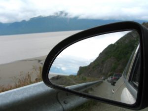 Traffic, mountains, and sea from side-view mirror of car.