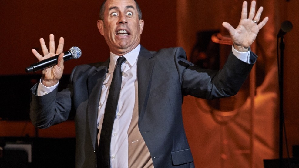 comedian Jerry Seinfeld holding a microphone with hands up and eyes wide in surprise