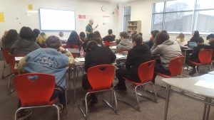 a photo from the back of a classroom shows a mix of student-engagement levels as the teacher speaks from the front