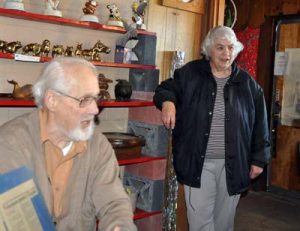 An elderly man sits in front of a display shelf with an elderly woman standing in the background.