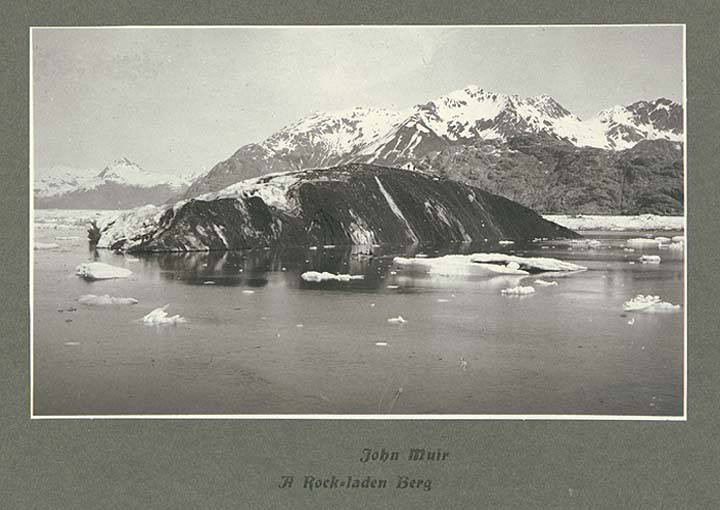 Black and white photograph of a gravel-covered iceberg floating in a glacial lake in Glacier Bay, Alaska.